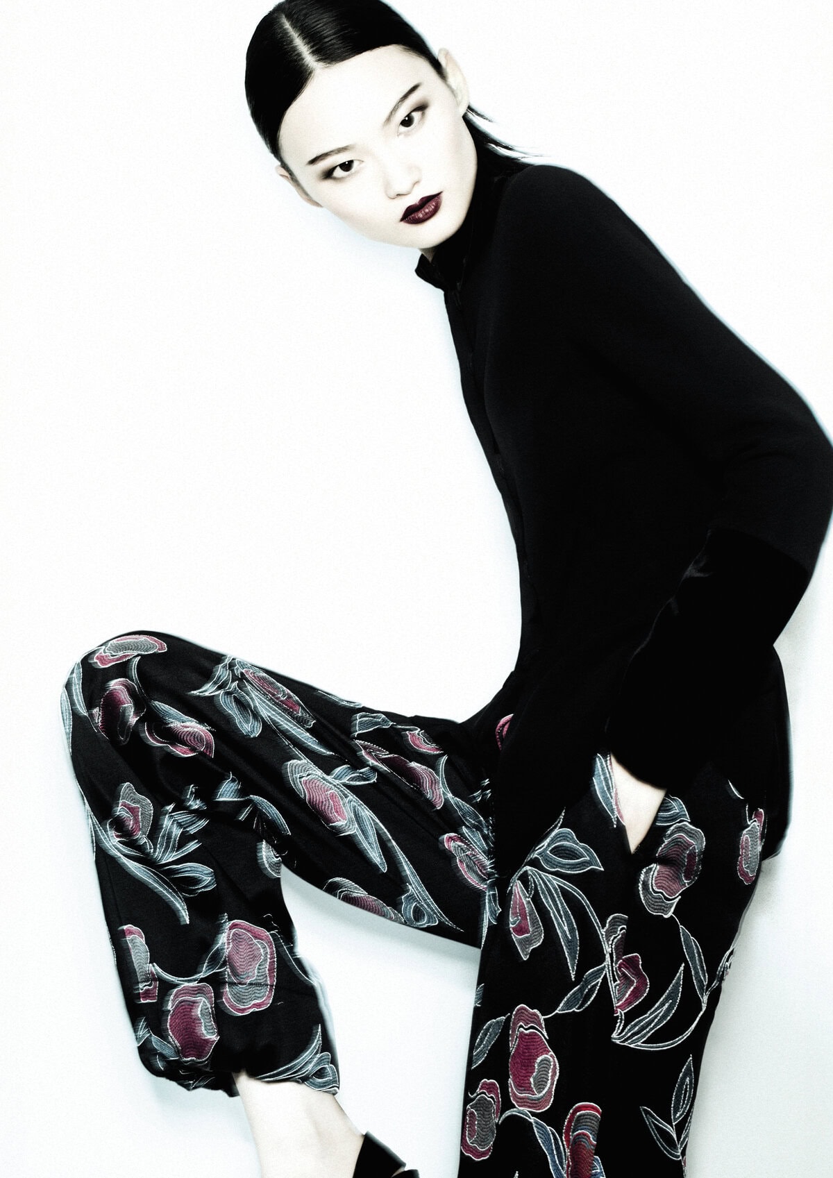 Model wearing a black top and floral patterned trousers against a clean white seamless studio background.