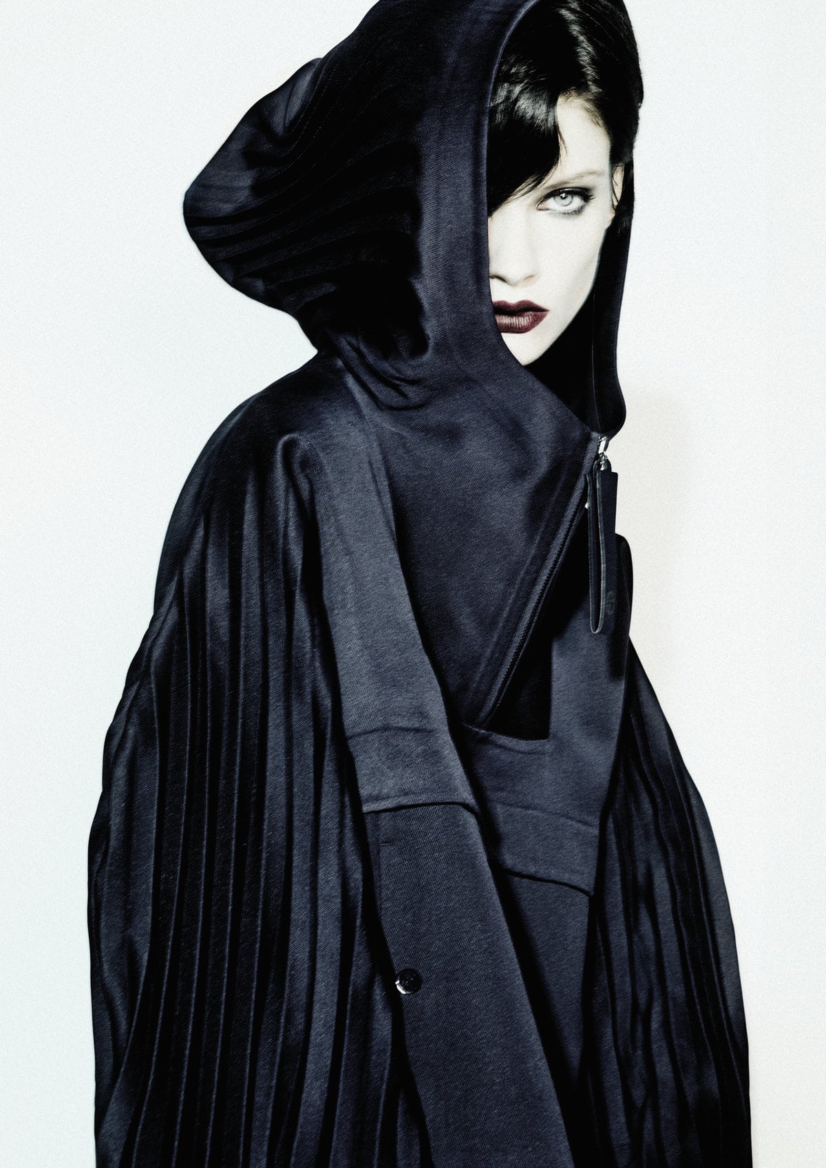 Model wearing a dark hooded garment with pleated details against a plain white background