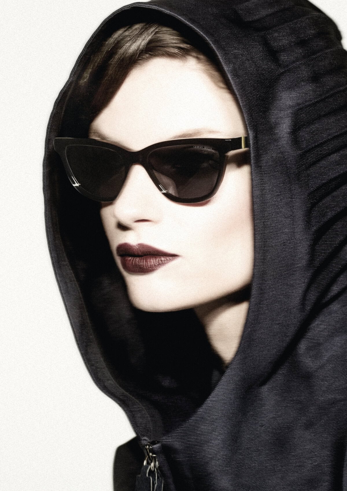 Model wearing dark sunglasses and a black hooded jacket against a white background for Giorgio Armani Fall 2024