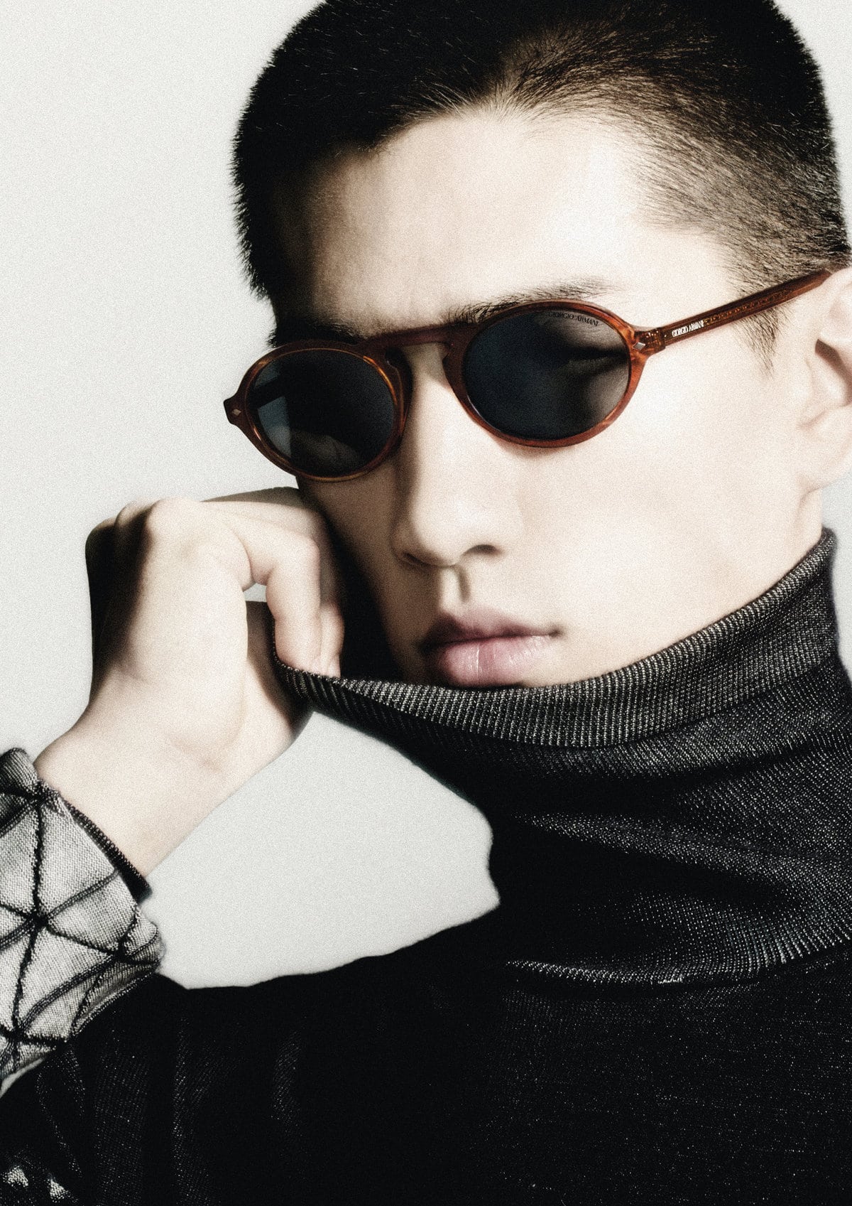 Close-up portrait of a man wearing dark round sunglasses and a high-neck dark sweater against a neutral background.