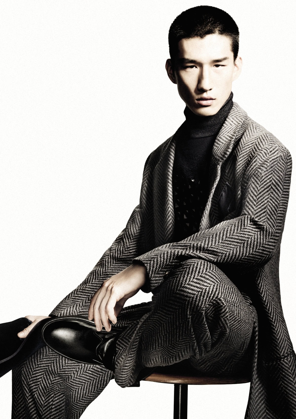 Man wearing a grey patterned suit and dark turtleneck sitting on a stool against a white background
