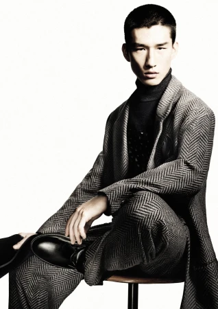 Man wearing a grey patterned suit and dark turtleneck sitting on a stool against a white background