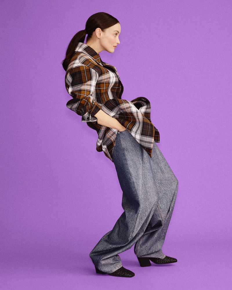A young adult woman poses in a plaid jacket and loose blue jeans against a light purple backdrop in a studio fashion shot.