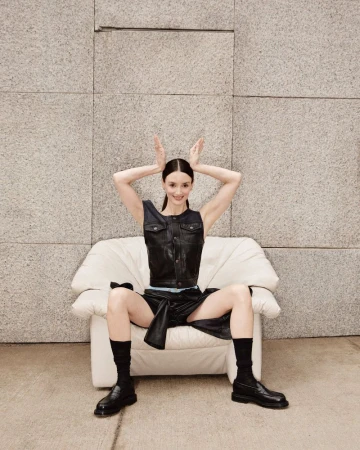 A woman in a black leather vest and skirt poses in a white armchair against a stone wall, making bunny ears with her hands.