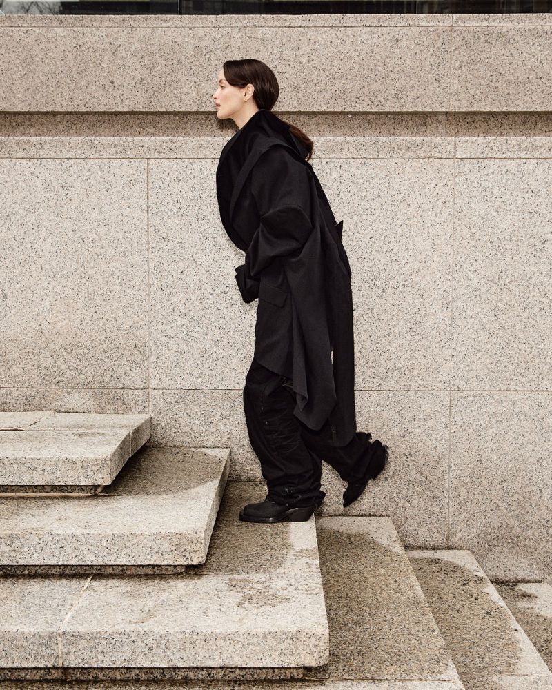 Woman in a dark coat ascends grey stone stairs outdoors, captured in a fashion editorial style with a shallow depth of field.