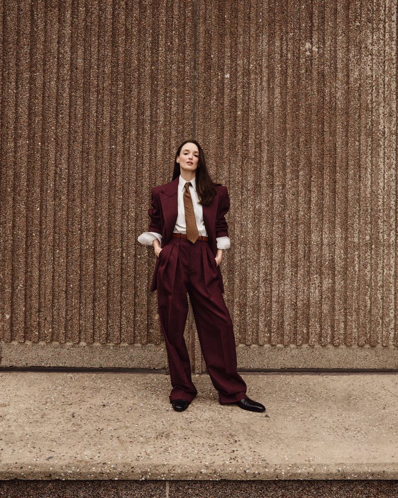 A woman in a burgundy suit stands confidently against a textured brown wall in an outdoor setting, casting a modern, fashion-forward look.