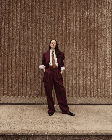 A woman in a burgundy suit stands confidently against a textured brown wall in an outdoor setting, casting a modern, fashion-forward look.