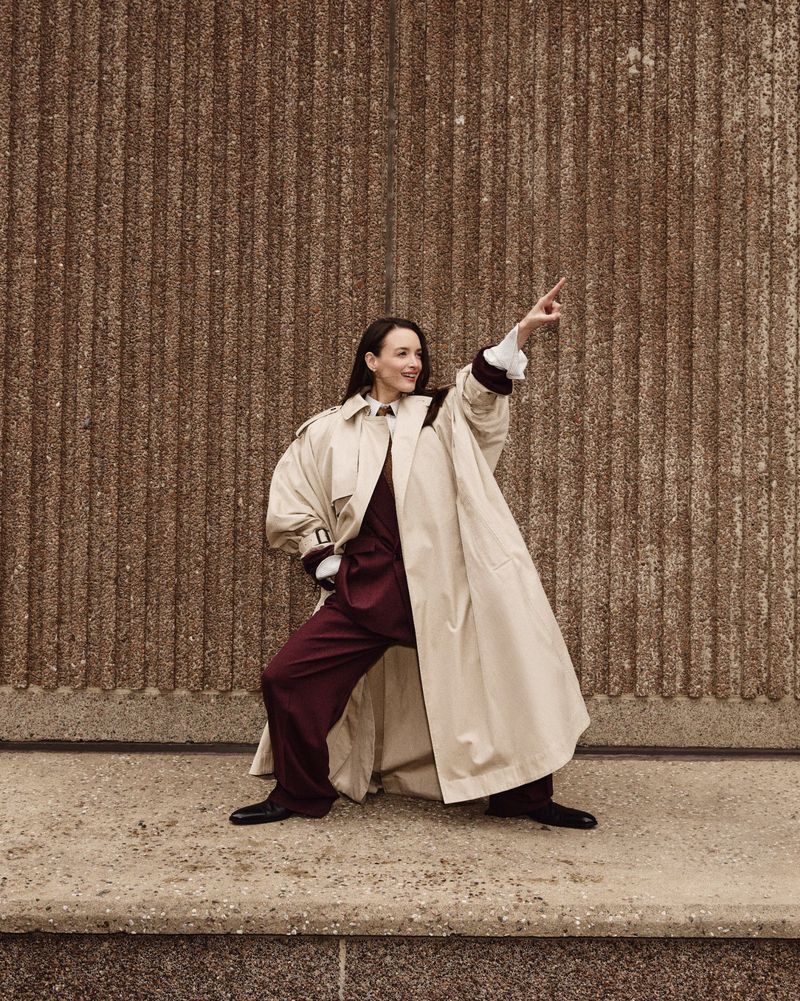 Woman in beige trench coat and maroon pants points up in front of textured concrete wall, creating a striking fashion portrait.