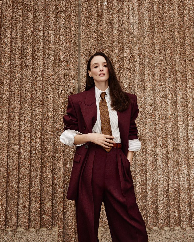 A woman in a burgundy suit stands indoors against a brown patterned wall, looking at the camera with a neutral expression and confident pose.