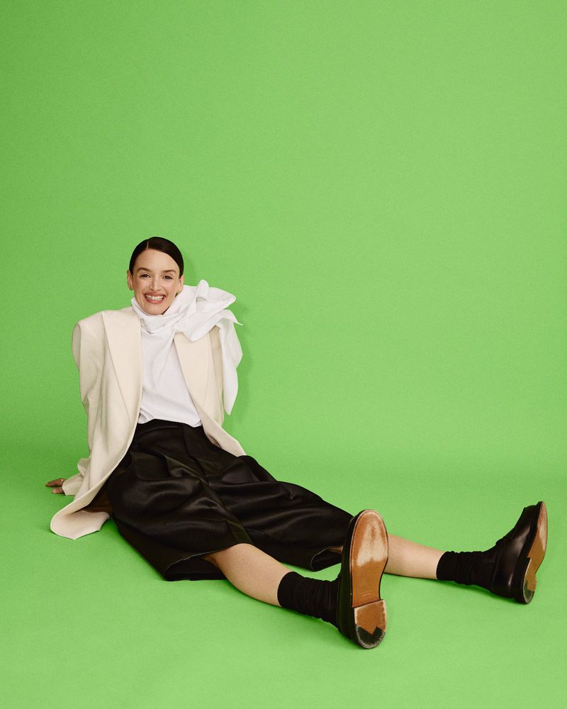 Smiling woman in a blazer and black culottes sitting on a green background, facing forward with legs extended towards the viewer.