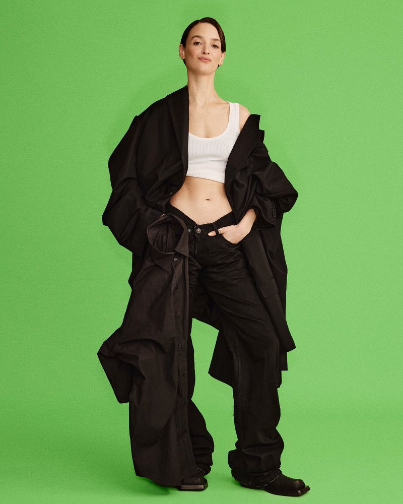 Full-length shot of a woman in a white top, black jacket, and baggy black pants against a green background in a studio setting.