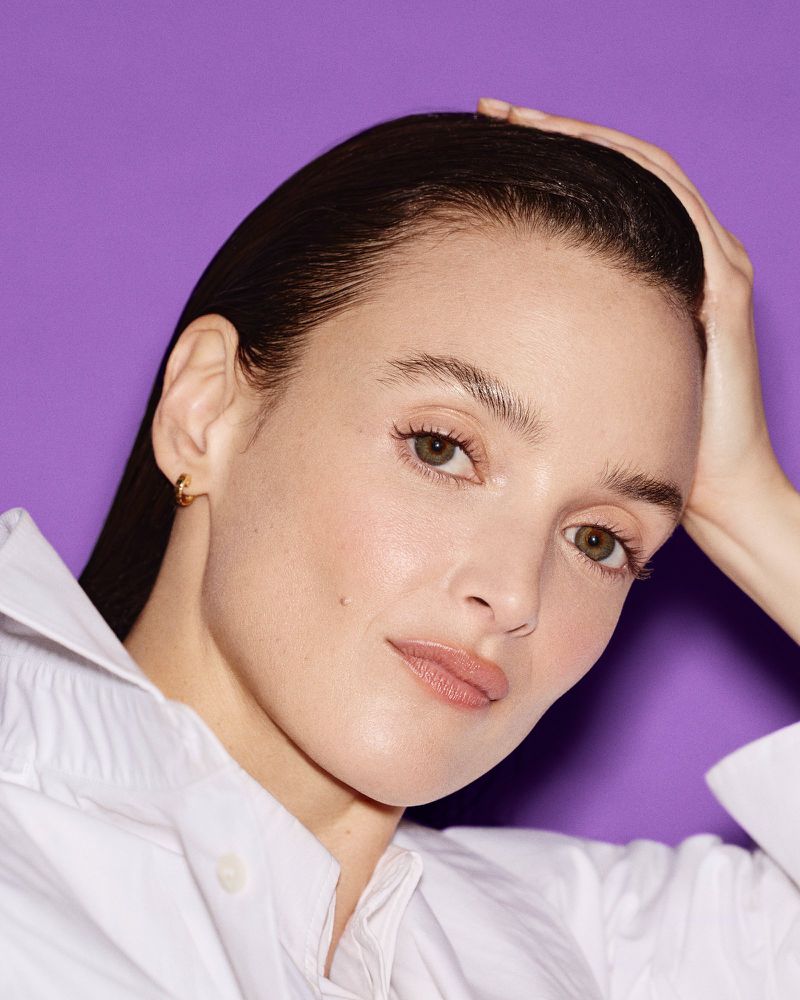 Close-up portrait of a young adult woman with wet-look hair, wearing a white shirt against a solid light purple background.