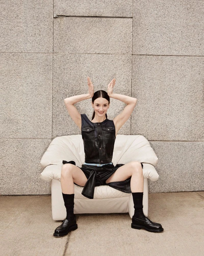 Charlotte Le Bon seated in a white armchair wearing a black leather vest and shorts against a stone wall background