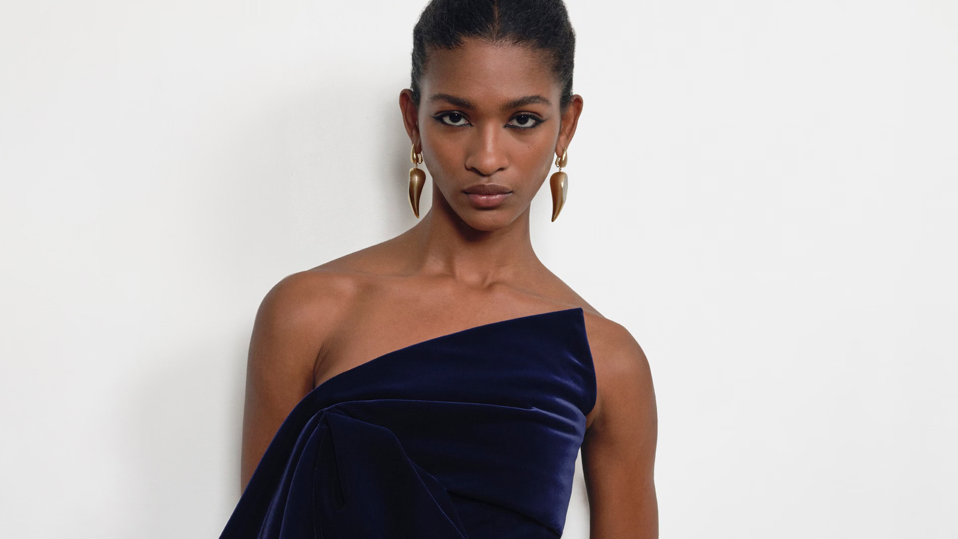 Model wearing a dark blue velvet one-shoulder top and sculptural gold earrings against a plain white background.