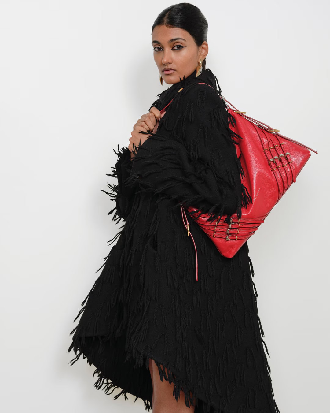 Model in black fringe coat carrying a large red leather handbag against a plain white studio background.