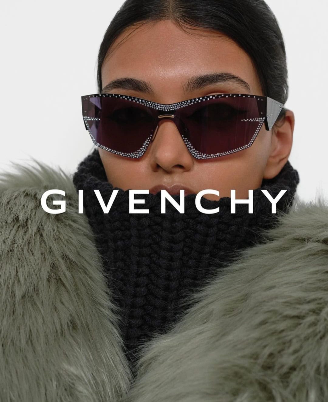 Close-up portrait of a model wearing crystal-embellished sunglasses and a black turtleneck under a furry green coat against a white background.