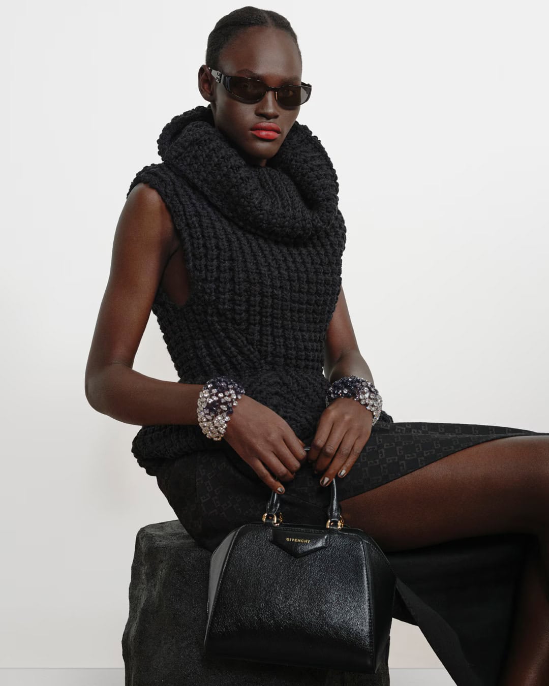 Model in a black textured knit top and sunglasses holding a black handbag against a light studio backdrop