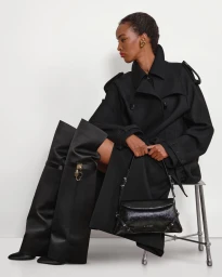 Model wearing a black coat and tall boots seated on a stool holding a handbag against a seamless white background.