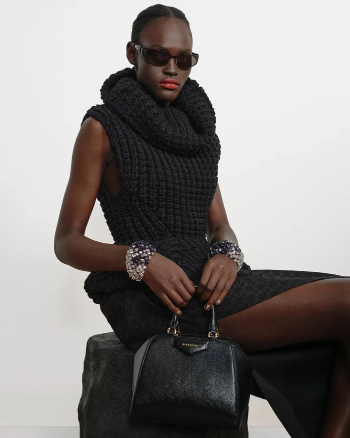 Model in a black textured knit top and sunglasses holding a black handbag against a light studio backdrop