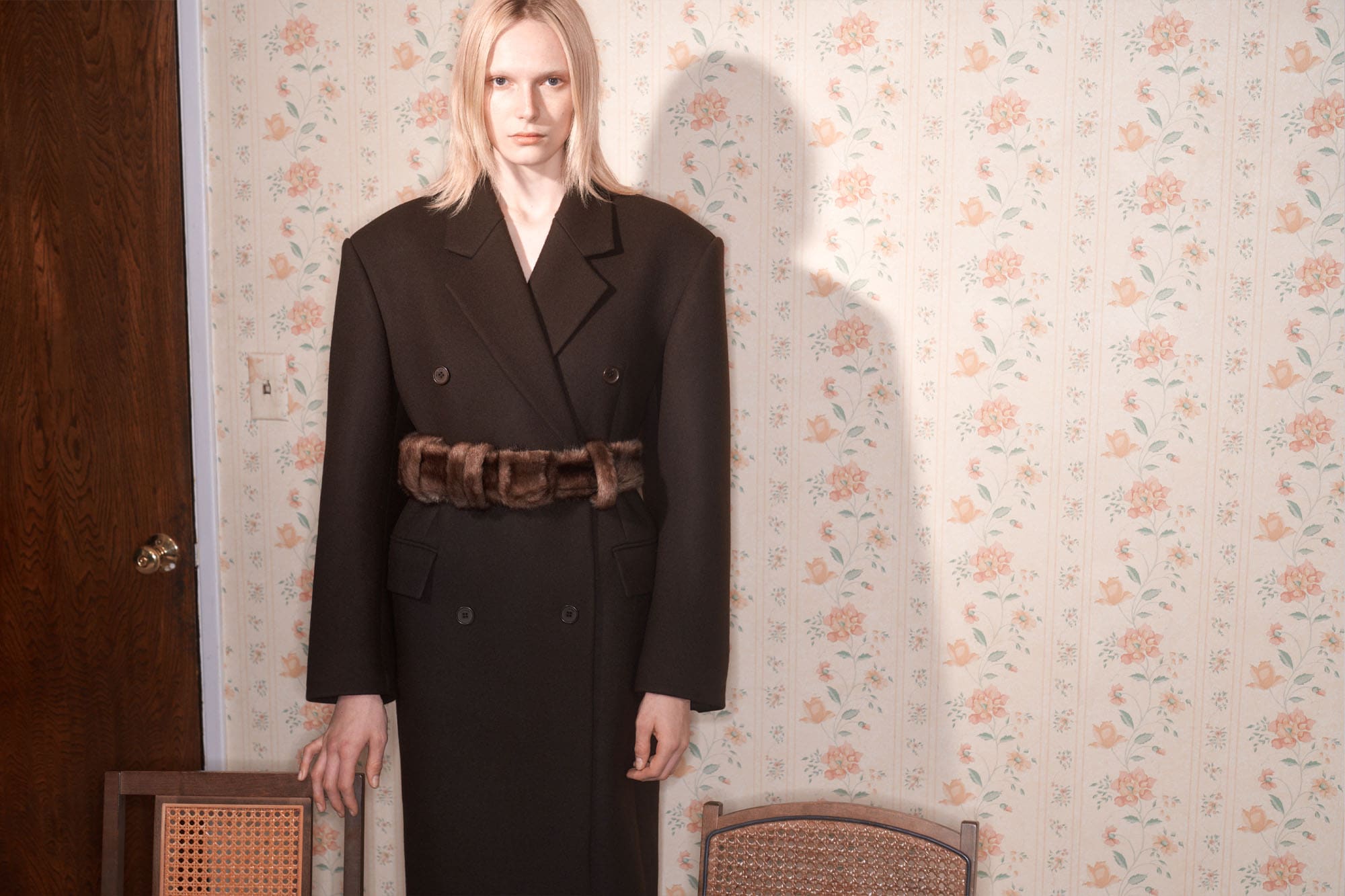 Model with long blonde hair wearing a dark oversized double-breasted coat with a thick textured belt against vintage floral wallpaper.