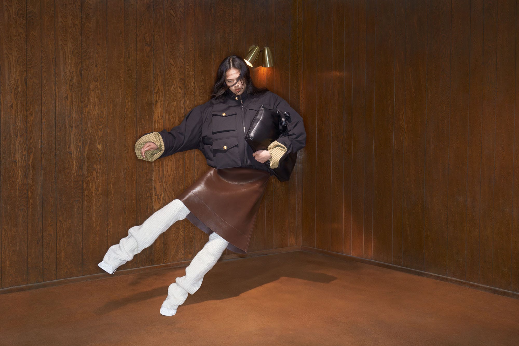 Model wearing dark jacket, brown skirt, and white leg warmers holding a black bag inside a wood-paneled room