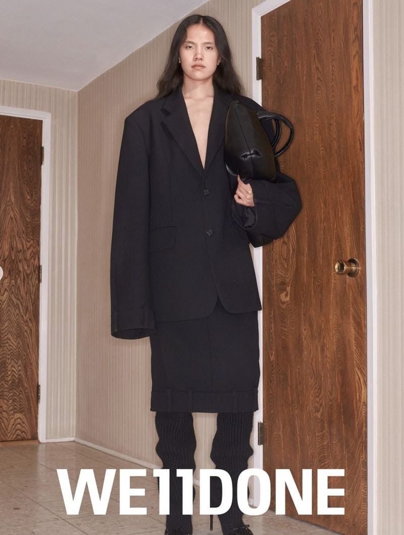 Model in oversized black blazer and skirt holding a black handbag inside a hallway with wooden doors