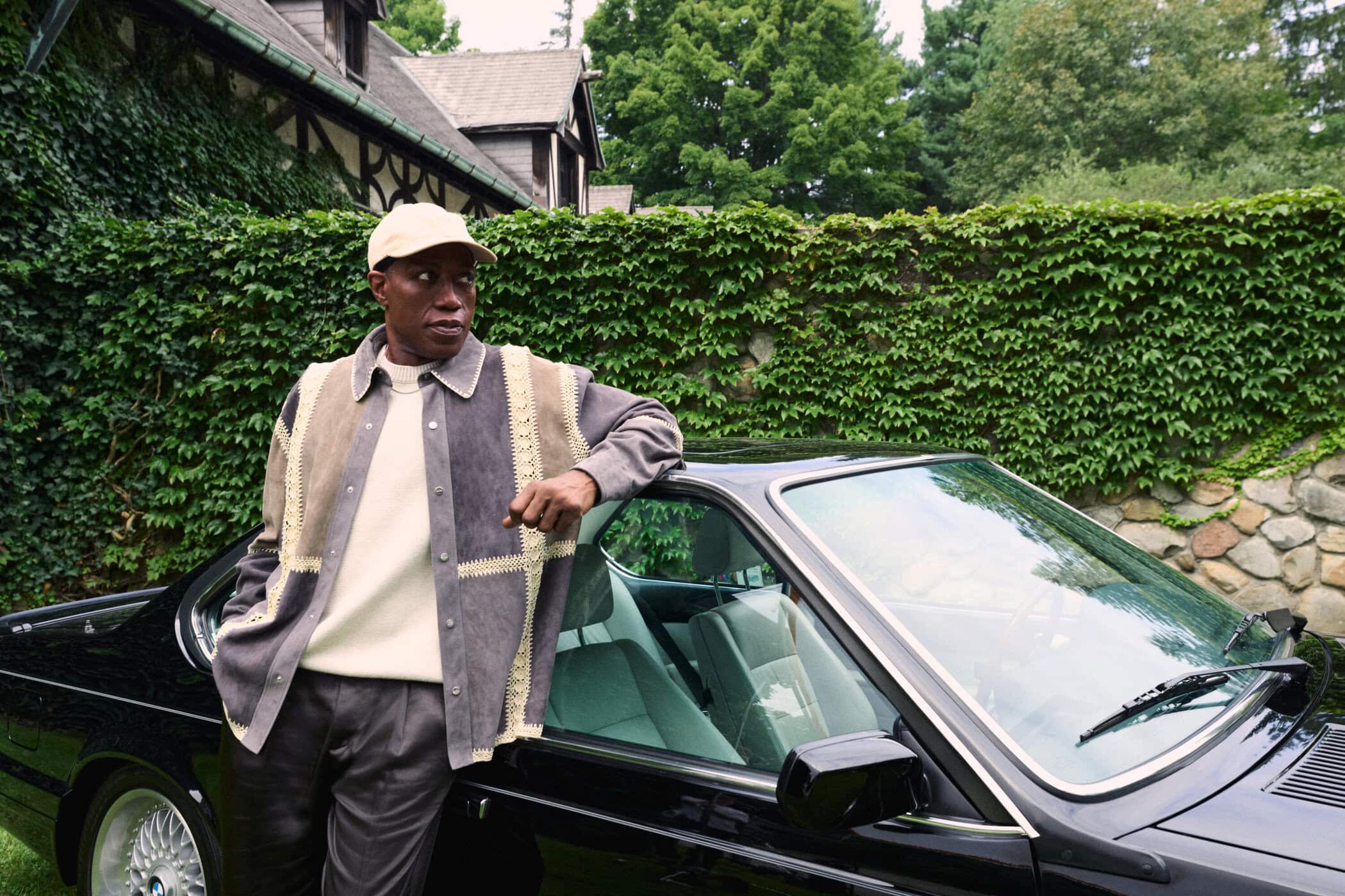 Wesley Snipes leaning against a vintage black car wearing a grey patchwork jacket and a beige cap