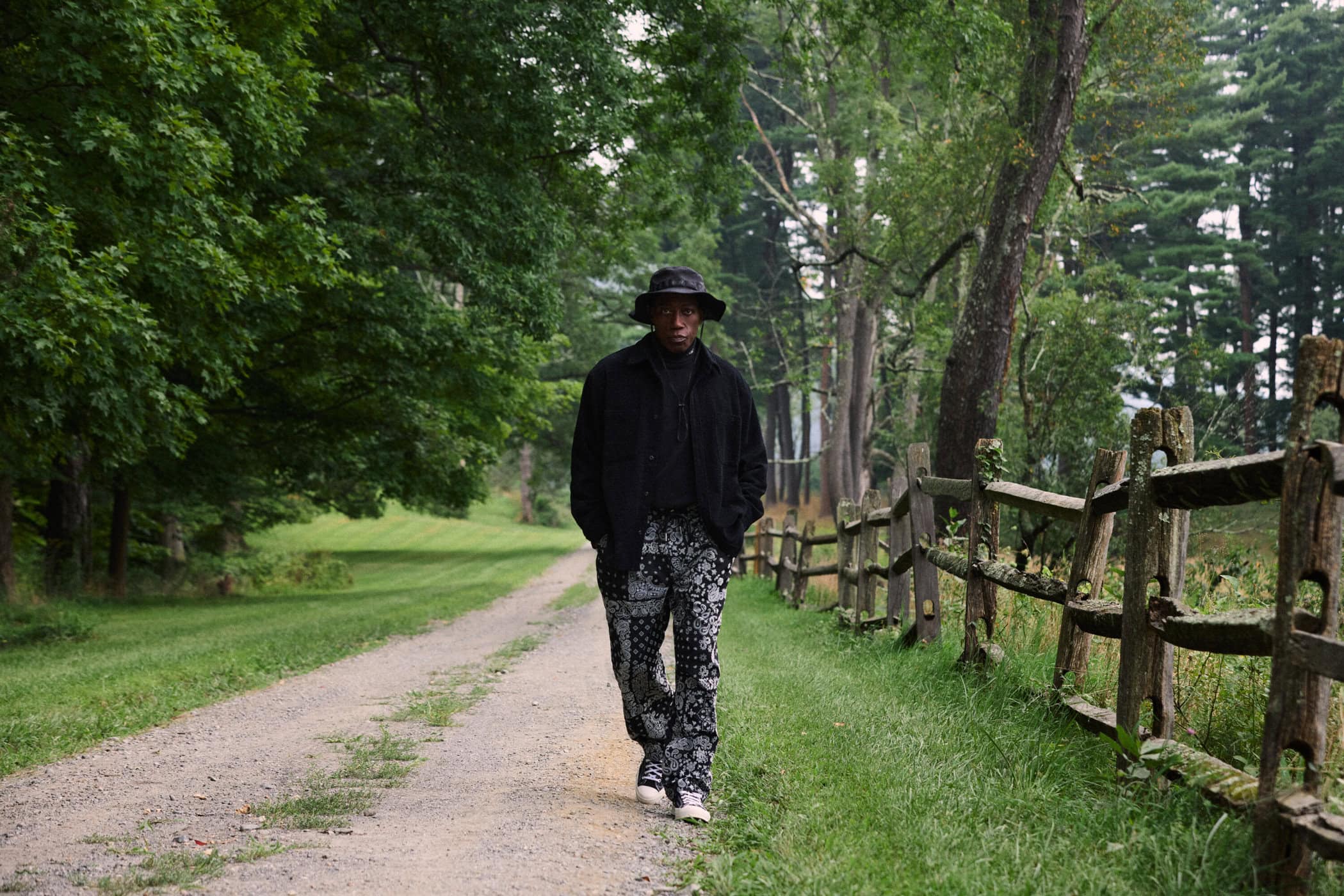 Wesley Snipes walking along a rural dirt path flanked by a rustic wooden fence and lush trees for Kith Fall 2024 Ad Campaign.