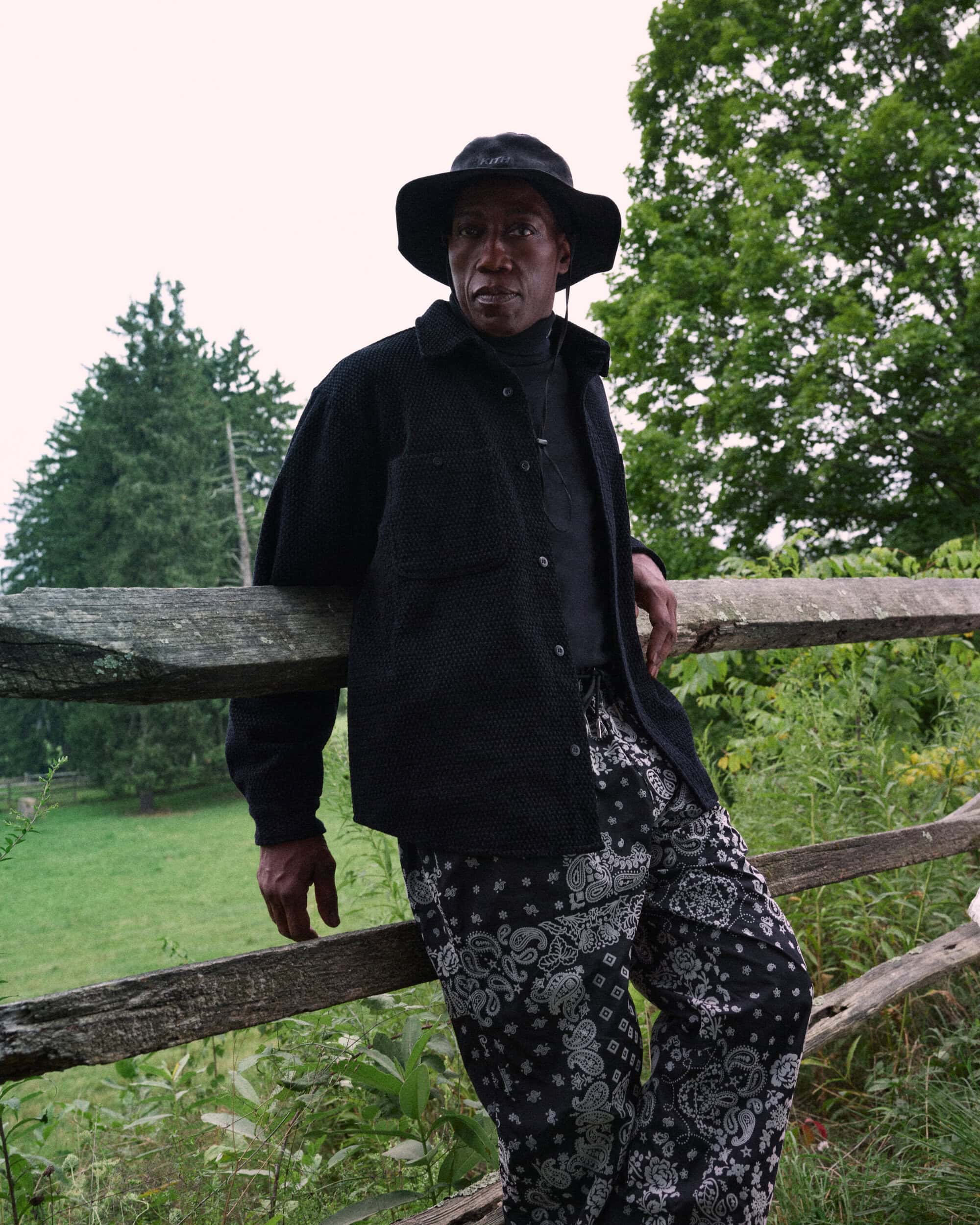Wesley Snipes wearing a black bucket hat and patterned trousers leaning against a wooden fence in a lush green outdoor setting.