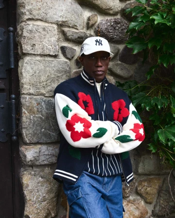 Wesley Snipes wearing a white baseball cap, floral bomber jacket, and denim jeans against a stone wall.