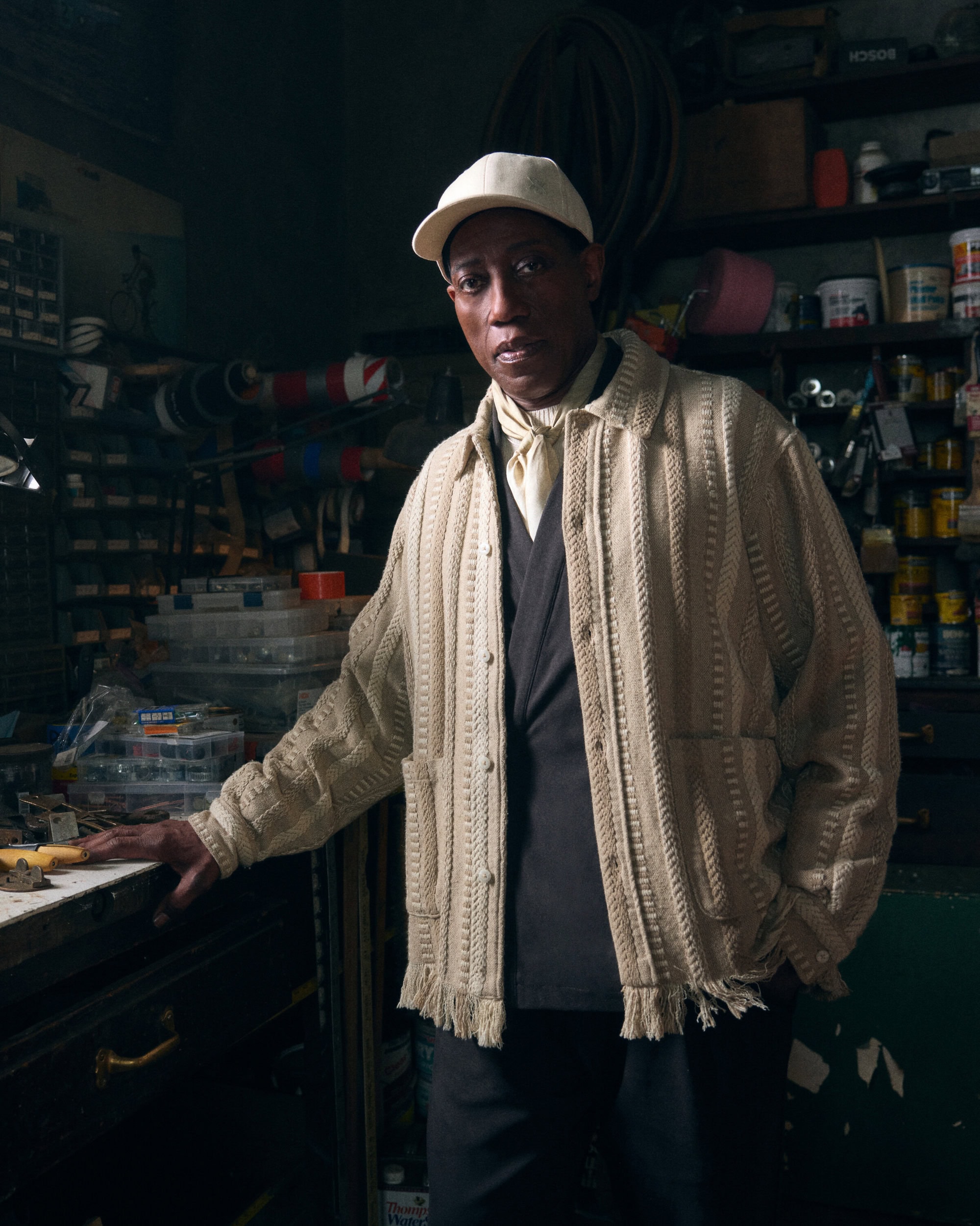 Wesley Snipes wearing a cream cable-knit cardigan and baseball cap standing in a cluttered workshop for Kith Fall 2024 campaign.