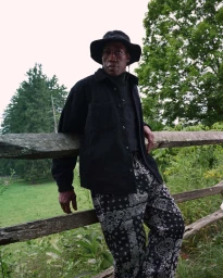 Wesley Snipes wearing a black bucket hat and patterned trousers leaning against a wooden fence in a lush green outdoor setting.