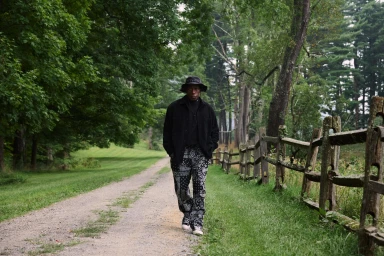 Wesley Snipes walking along a rural dirt path flanked by a rustic wooden fence and lush trees for Kith Fall 2024 Ad Campaign.