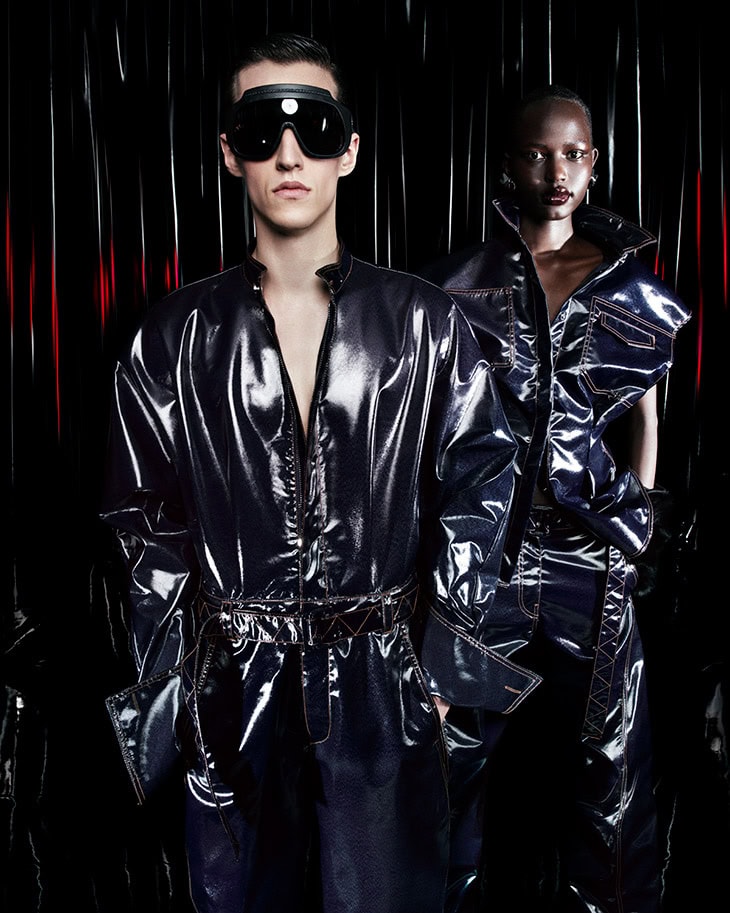 Two models wearing glossy black leather outfits and dark futuristic visors against a dark reflective background.