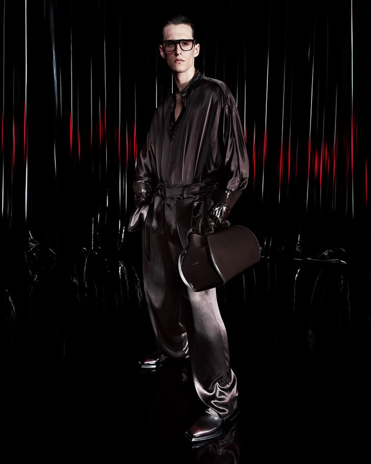 Model wearing dark silky shirt and trousers holding a cylindrical dark handbag against a dark backdrop with vertical red accents.