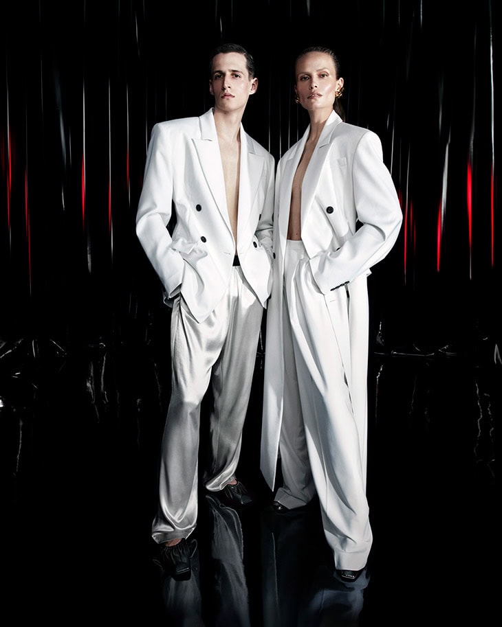 Two models wearing matching white tailored suits standing against a dark reflective background for Ferrari.