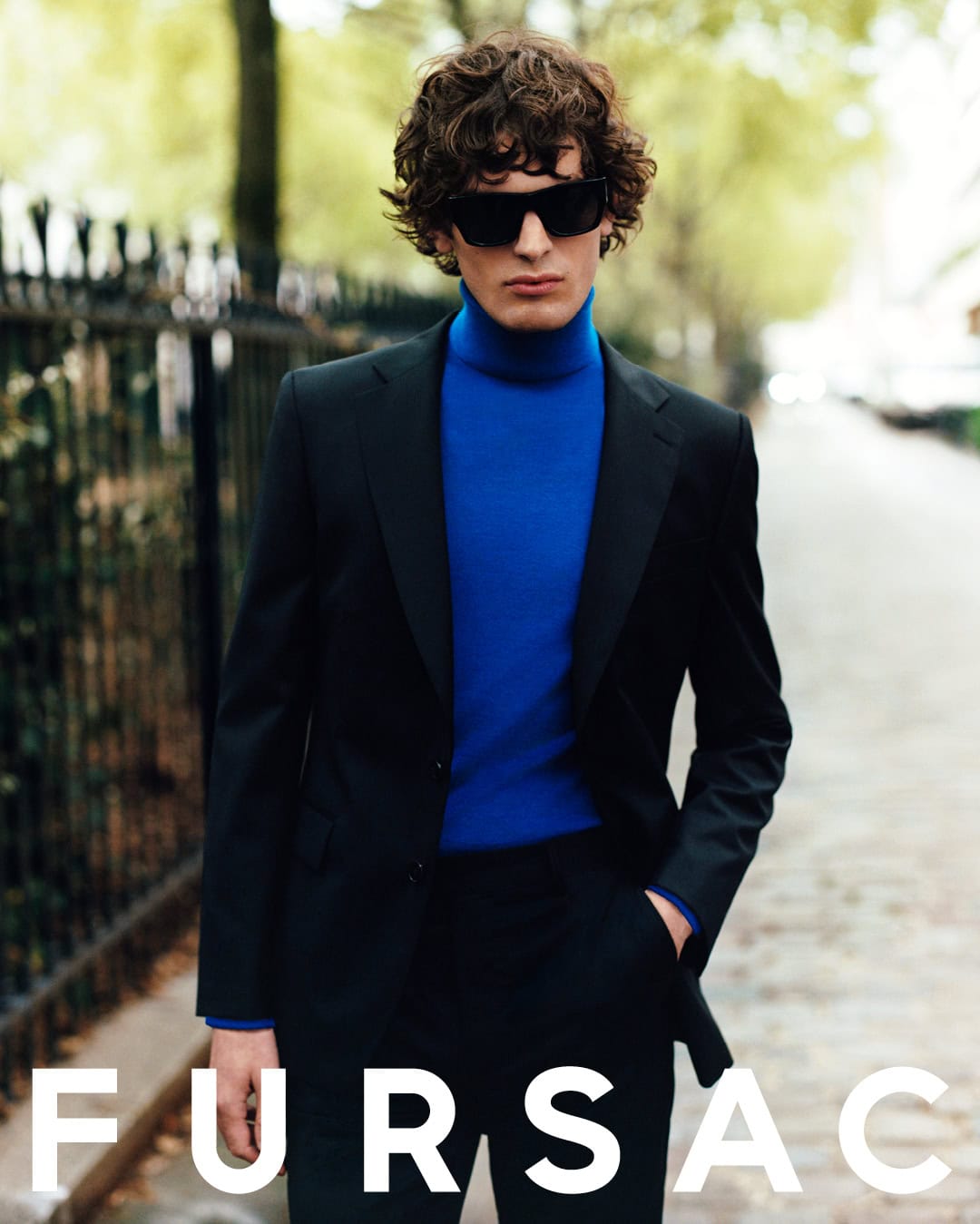 Model wearing dark sunglasses, a black suit, and a vibrant blue turtleneck outdoors on a paved street by a black metal fence.