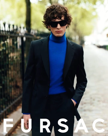 Model wearing dark sunglasses, a black suit, and a vibrant blue turtleneck outdoors on a paved street by a black metal fence.