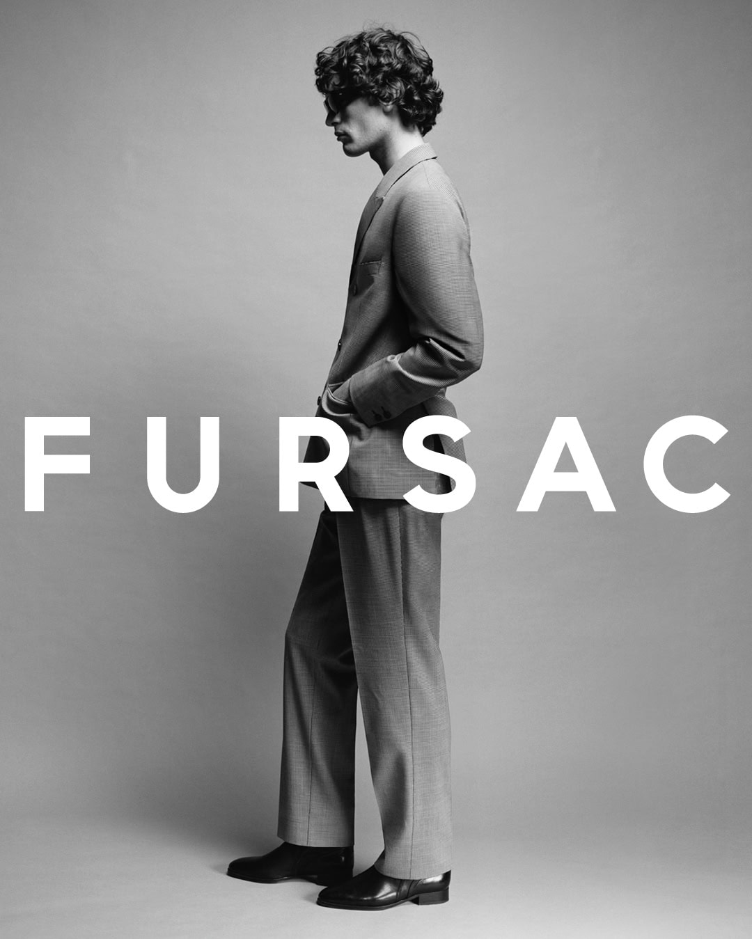 Profile view of male model in tailored grey suit standing against a plain grey background with bold white text overlay