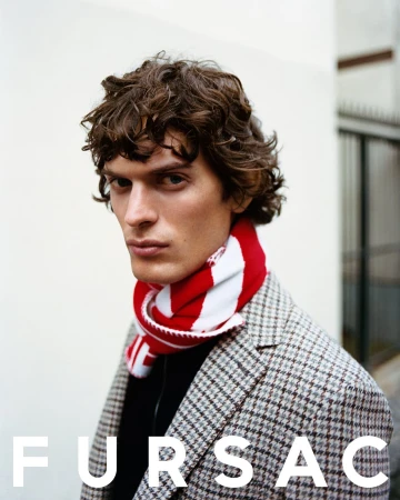 Portrait of a male model wearing a patterned blazer and a red and white scarf against a light architectural background.