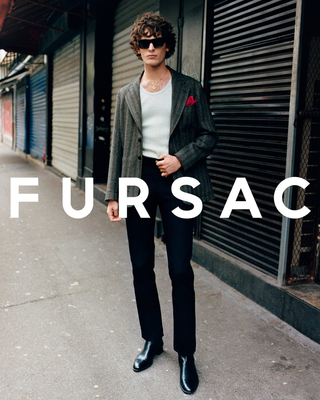 Model standing on a city sidewalk wearing a grey patterned blazer over a white tank top with dark trousers and black boots