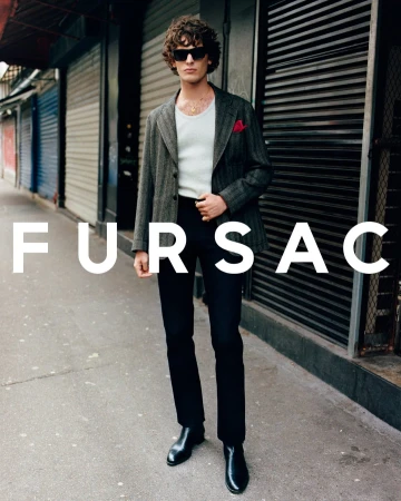 Model standing on a city sidewalk wearing a grey patterned blazer over a white tank top with dark trousers and black boots