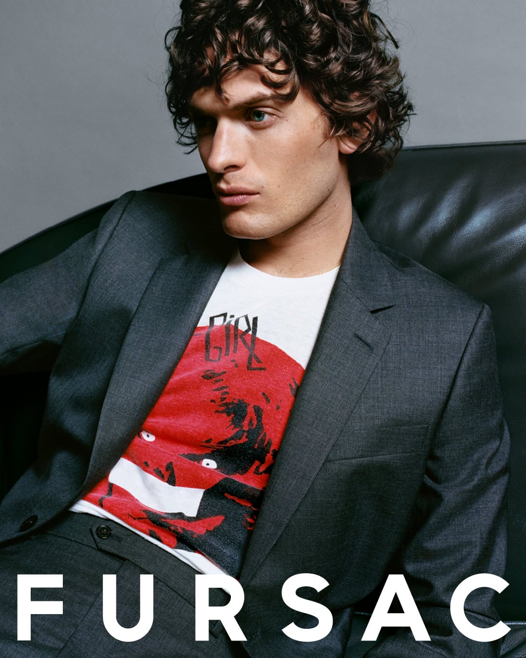 Male model with curly dark hair wearing a grey suit jacket and graphic t-shirt seated against a grey backdrop