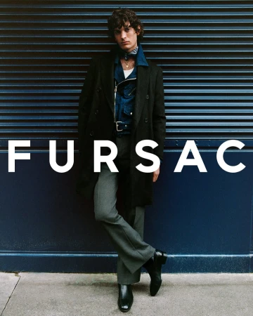 Male model wearing a dark coat and trousers leaning against a dark blue rolling shutter door with brand text