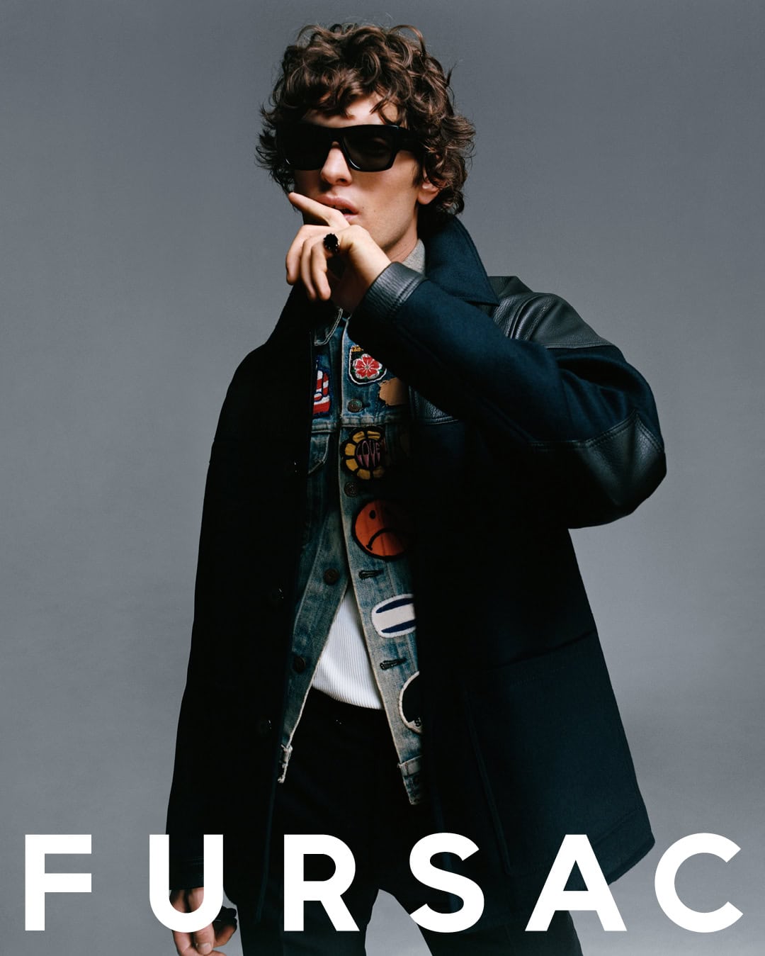 Model wearing dark sunglasses, patch-covered denim jacket, and dark coat with finger to lips against a grey background
