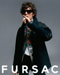 Model wearing dark sunglasses, patch-covered denim jacket, and dark coat with finger to lips against a grey background