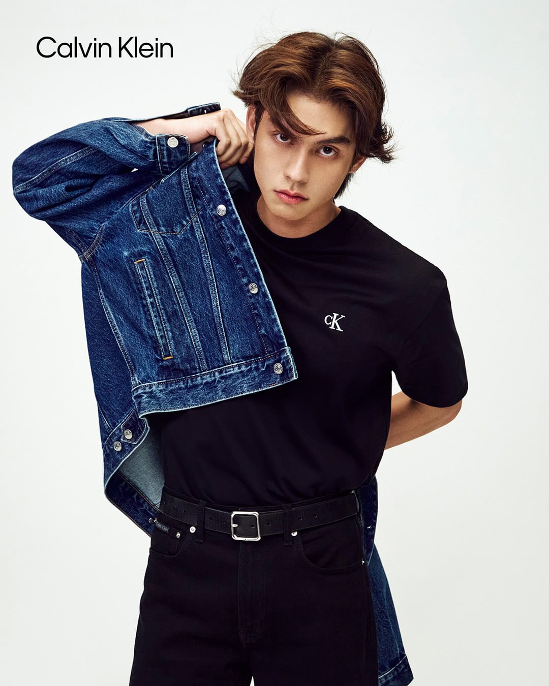 Model Bright wearing a black t-shirt and dark jeans holding a denim jacket against a white background for Calvin Klein