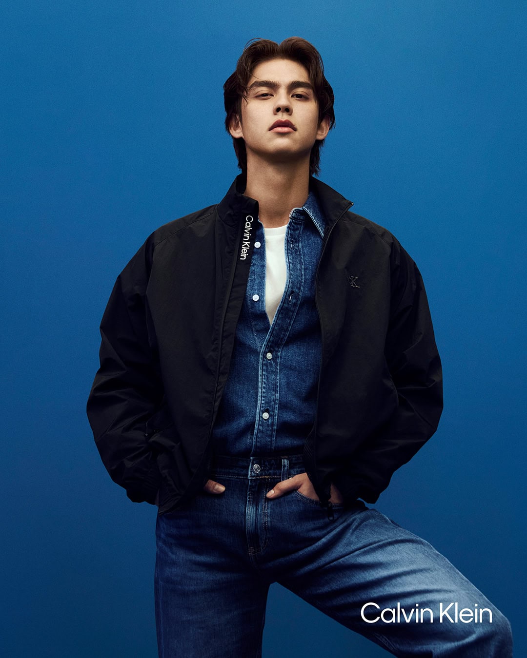 Model Bright wearing a black jacket and denim shirt posing against a solid blue background for Calvin Klein