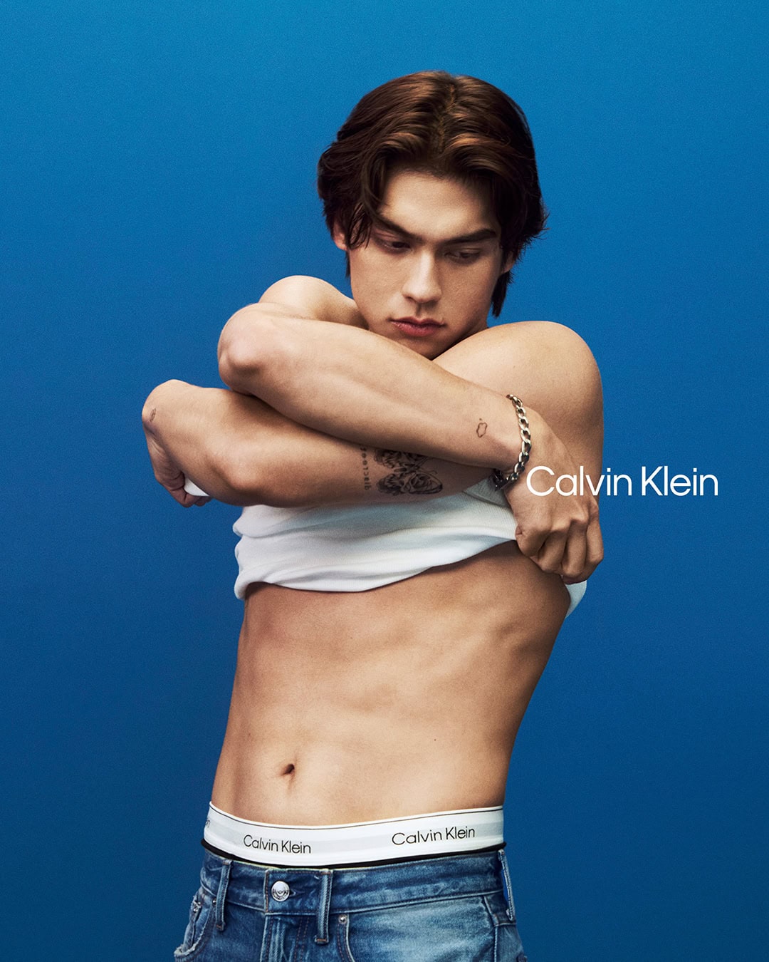 Man taking off a white t shirt against a solid blue background wearing Calvin Klein denim jeans