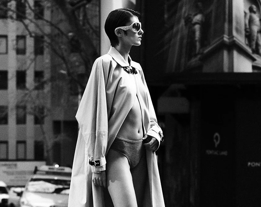 Monochrome fashion photograph of a model in sunglasses and an oversized coat standing outdoors on a city street.